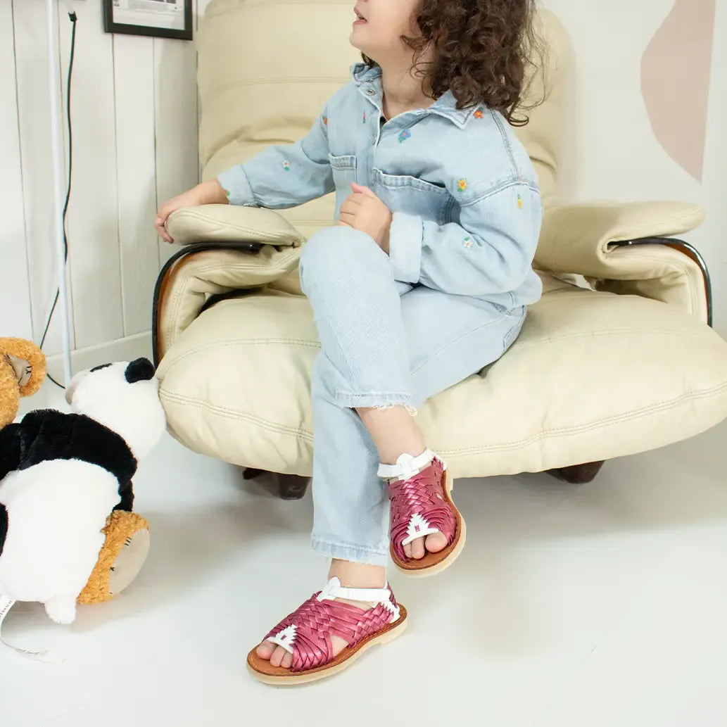 Une petite fille assise sur un fauteuil beige porte les Sandales Sandalita Rose Flashy Blanc. Elle croise les jambes et regarde sur le côté.