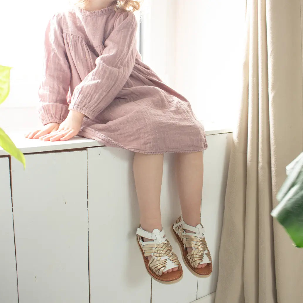 Une petite fille assise sur un rebord de fenêtre porte les Sandales Sandalita Champagne Blanc ainsi qu'une robe en live rose poudrée.